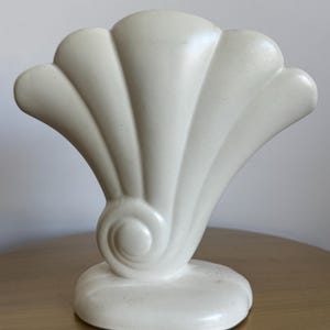 May include: A cream-colored, seashell-shaped decorative object. The sculpture features a fan-like design with curved ridges and a spiral detail at the base. The base is oval-shaped. The object is made of a solid material.