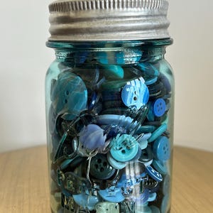 May include: A blue-tinted glass Mason jar with a silver metal lid, filled with a collection of assorted buttons in various shades of blue, teal, and navy. The glass jar features the embossed text "Perfect Mason" on the front surface.