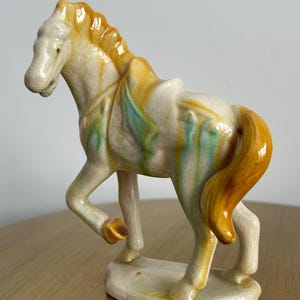 May include: Ceramic horse figurine in a standing pose. The horse is primarily cream-colored with yellow, orange, and green accents. The mane and tail are a vibrant orange. The figurine has a textured, crackled glaze.