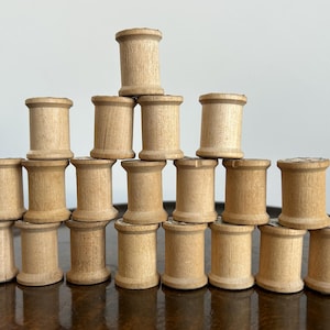 May include: A collection of twenty-one vintage wooden spools arranged in a pyramid shape. The spools are cylindrical with a slightly flared base and top, and are made of light-colored wood. The arrangement is on a dark brown surface.