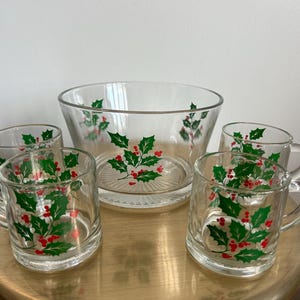May include: A clear glass punch bowl and matching mugs set. The bowl and mugs are decorated with a green holly leaf and red berry design. The set includes a large bowl and five mugs with handles.
