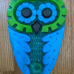 May include: A wooden owl carving with a vibrant color scheme. The owl features a green head with gear-like details, blue flower-shaped eyes, and blue wings. The body is a mix of blue and green, creating a whimsical and decorative piece.