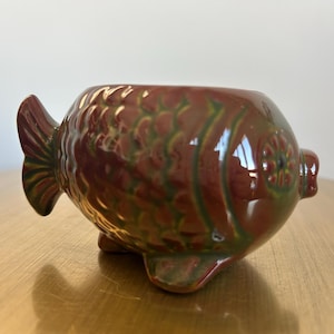 May include: A ceramic fish-shaped bowl with a glossy finish. The bowl is primarily reddish-brown with green accents, and features a detailed fin and tail design. The bowl is resting on a wooden surface.