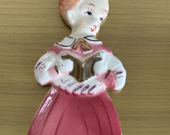 Könnte beinhalten: Vintage Weihnachtsornament aus Keramik in Form eines Chorknaben. Das Ornament hat ein rotes und weißes Gewand mit einem goldenen Buch. Der Chorknabe hat rosige Wangen, blaue Augen und einen roten Mund. Zum Aufhängen ist eine goldene Schnur befestigt.