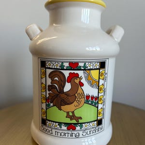 May include: A white ceramic milk jug with a yellow rim and two small handles. The jug features a colorful illustration of a rooster, sun, and flowers, with the text "Good morning Sunshine".