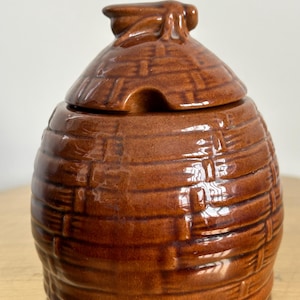 May include: A brown ceramic honey pot with a lid. The pot has a textured, woven basket design. The lid has a small handle and a notch for a spoon. The honey pot is sitting on a gold surface.