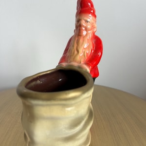 May include: A ceramic figurine of a gnome wearing a bright red hat and jacket, standing behind a large, cream-colored sack with a gold-toned rim. The gnome has a long, light-colored beard and is positioned as if pushing the sack forward.