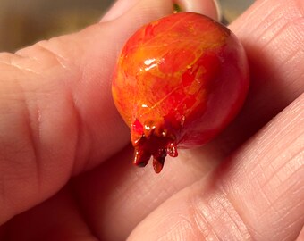 May include: A small, round, red and orange decorative pomegranate with a textured surface. The pomegranate has a brown stem and a detailed, textured crown. The fruit is held in a hand, showcasing its size and color.