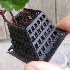 Puede incluir: Una pequeña maceta negra impresa en 3D con un diseño de celosía, que contiene tierra y una pequeña planta con hojas verdes y blancas. La maceta está sostenida por una mano, mostrando su estructura arquitectónica única.