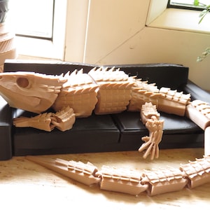 Könnte beinhalten: Ein beigefarbenes, artikuliertes, 3D-gedrucktes Modell einer Eidechse, das auf einem schwarzen Sofa liegt. Die Eidechse hat einen detaillierten, segmentierten Körper und einen stacheligen Rücken.