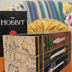 May include: A stack of three decorative wooden boxes with illustrations from The Lord of the Rings. The boxes feature maps, swords, and a tree design. A copy of "The Hobbit" book is visible in the background.