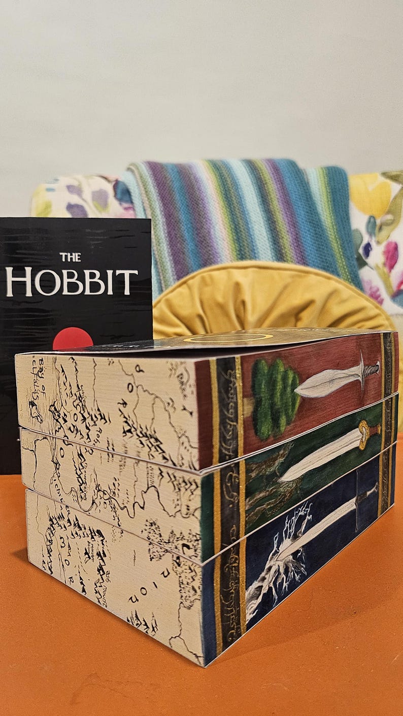 May include: A stack of three decorative wooden boxes with illustrations from The Lord of the Rings. The boxes feature maps, swords, and a tree design. A copy of "The Hobbit" book is visible in the background.