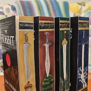 May include: A collection of J.R.R. Tolkien books, including "The Hobbit," with custom painted spines featuring swords and fantasy imagery. The books are displayed on an orange surface.