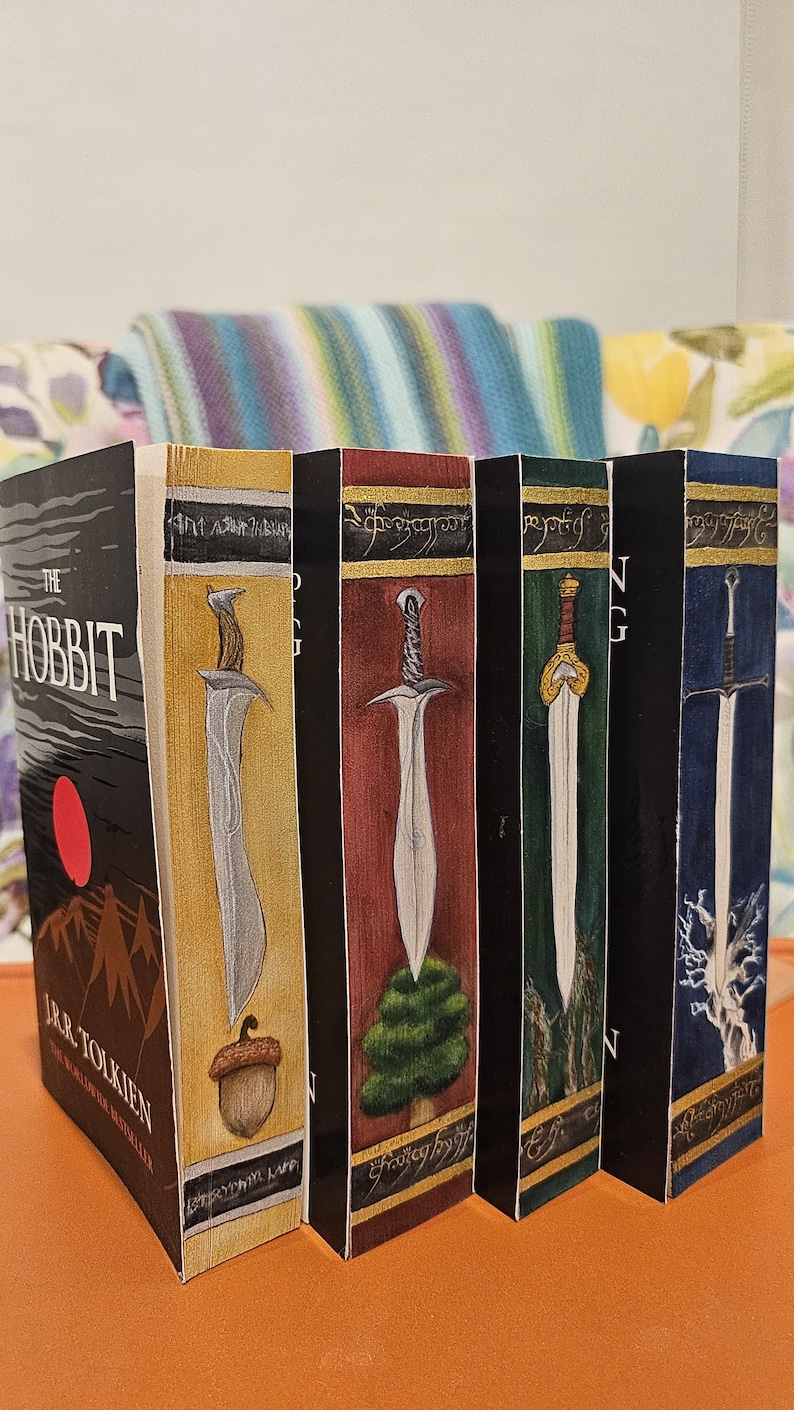 May include: A collection of J.R.R. Tolkien books, including "The Hobbit," with custom painted spines featuring swords and fantasy imagery. The books are displayed on an orange surface.