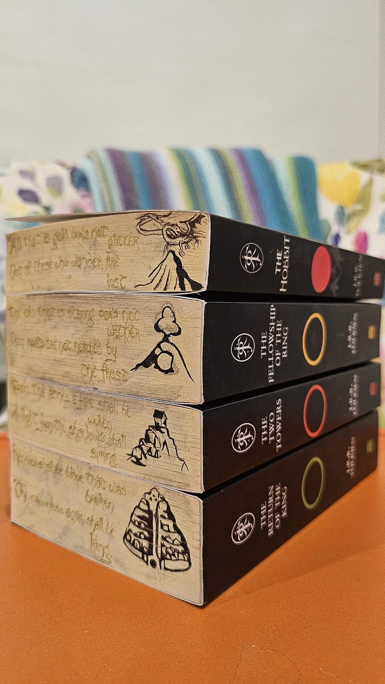 May include: A stack of four books from the Lord of the Rings series. The books have black spines with titles and colored circles. The top of each book has a wood-grain design with handwritten text and illustrations.