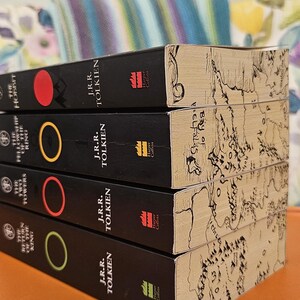 May include: A stack of four books by J.R.R. Tolkien, including "The Hobbit" and "The Return of the King." The books have black spines with colored circles and the Harper Collins logo. The edges of the books feature a map design.