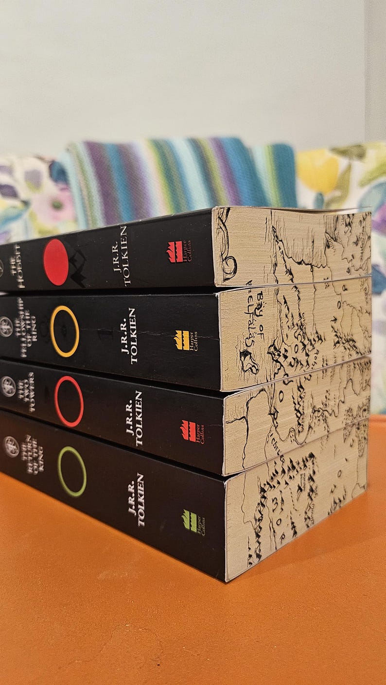 May include: A stack of four books by J.R.R. Tolkien, including "The Hobbit" and "The Return of the King." The books have black spines with colored circles and the Harper Collins logo. The edges of the books feature a map design.