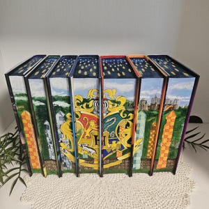 May include: A collection of seven hardcover books with custom painted covers. The spines feature a night sky design with stars. The covers display various crests and illustrations, including a castle and house emblems. The books are arranged on a white surface.
