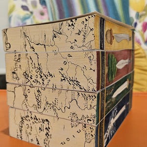 May include: A stack of four decorative boxes. The sides of the boxes feature a map design with black ink on a tan background. The top edges of the boxes have illustrations of a sword, a leaf, a spear, and a sword.
