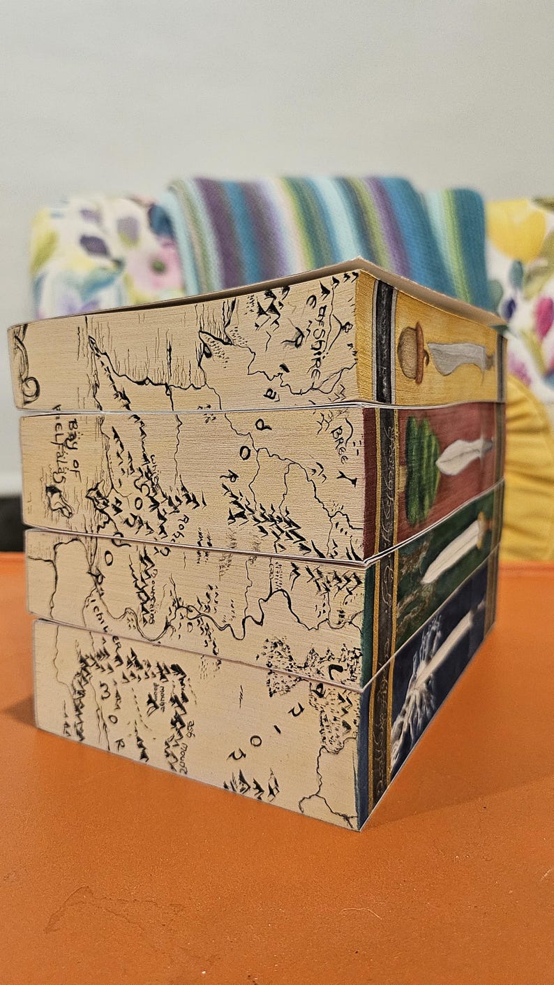 May include: A stack of four decorative boxes. The sides of the boxes feature a map design with black ink on a tan background. The top edges of the boxes have illustrations of a sword, a leaf, a spear, and a sword.