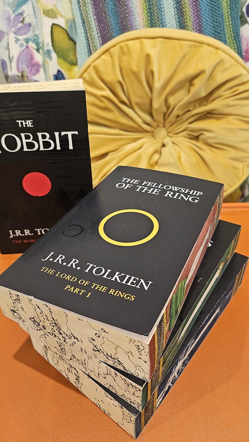 May include: Stack of J.R.R. Tolkien's "The Lord of the Rings" books, with "The Fellowship of the Ring" visible. The book covers are black with gold and yellow accents. "The Hobbit" book is also visible.
