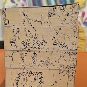 May include: A stack of four books with light brown covers. Each cover features a detailed black and white map design with text, including "Bay of Belfalas" and "The Shire". The books are stacked on top of each other.