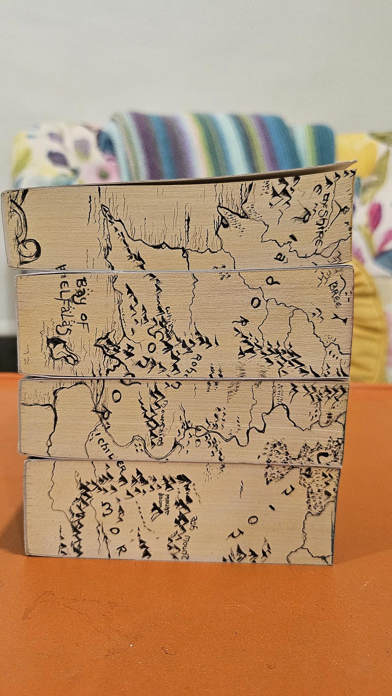 May include: A stack of four books with light brown covers. Each cover features a detailed black and white map design with text, including "Bay of Belfalas" and "The Shire". The books are stacked on top of each other.