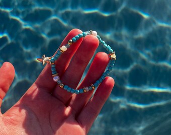 May include: A turquoise and white beaded bracelet with a gold starfish charm. The bracelet is held in a hand against a blue water background.