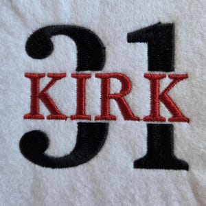 Op de afbeelding: Witte stof met geborduurde tekst en cijfers. Het nummer "31" is in zwart, met de naam "KIRK" geborduurd in rood. Het ontwerp is gecentreerd op de stof.