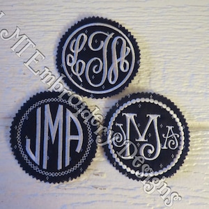 May include: Three embroidered fabric circles with white lettering on a dark blue background. The circles feature the initials "LJ", "JMA", and "MJA".