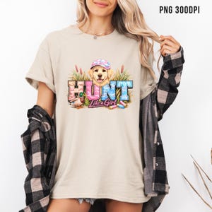 May include: Beige t-shirt with a graphic that says "HUNT Like a Girl." The graphic features a golden retriever wearing a hat, a duck, and colorful text. The shirt is a casual style.
