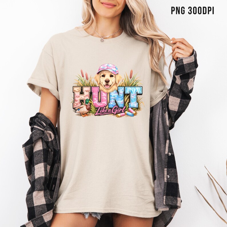May include: Beige t-shirt with a graphic that says "HUNT Like a Girl." The graphic features a golden retriever wearing a hat, a duck, and colorful text. The shirt is a casual style.