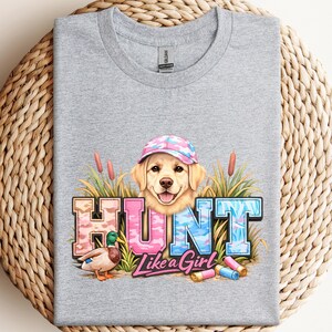 May include: A heather gray t-shirt featuring a golden retriever wearing a pink and blue camouflage cap. The shirt has the word "HUNT" in camouflage lettering, a duck, and the phrase "Like a Girl".