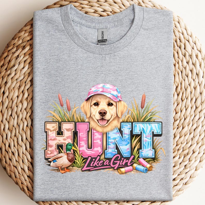 May include: A heather gray t-shirt featuring a golden retriever wearing a pink and blue camouflage cap. The shirt has the word "HUNT" in camouflage lettering, a duck, and the phrase "Like a Girl".