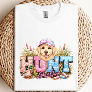 May include: White t-shirt featuring a graphic of a golden retriever wearing a pink and blue camouflage baseball cap. The word "HUNT" is in pink and blue camouflage lettering, with the phrase "Like a Girl" below. A duck and shotgun shells are also in the graphic.