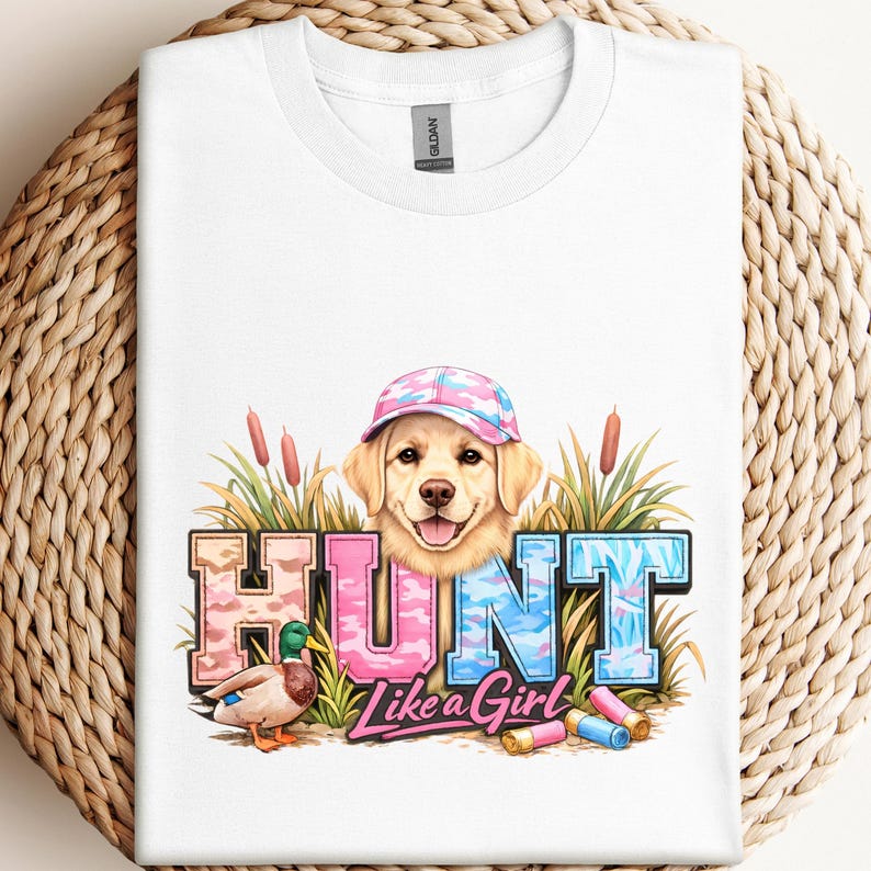 May include: White t-shirt featuring a graphic of a golden retriever wearing a pink and blue camouflage baseball cap. The word "HUNT" is in pink and blue camouflage lettering, with the phrase "Like a Girl" below. A duck and shotgun shells are also in the graphic.
