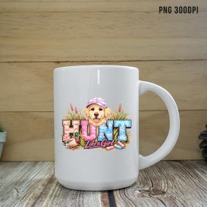 May include: White ceramic mug with a wraparound graphic. The graphic features a golden retriever wearing a camouflage hat, the word "HUNT" in pink and blue letters, and the phrase "Like a Girl". A duck and shotgun shells are also included.
