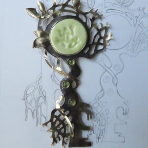 May include: A metal brooch shaped like a tree with a pale green circular center piece featuring embossed markings. The design includes metallic leaves, three small green inset stones along the trunk, and an intricate branch-like structure.