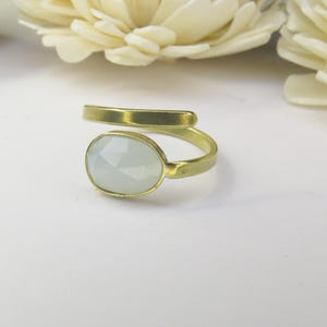 May include: A bypass ring featuring a matte gold-toned metal band and a single oval-shaped, faceted, pale translucent stone set in a bezel. The ring has an open design where the ends of the band do not meet, showcasing a smooth, metallic finish.