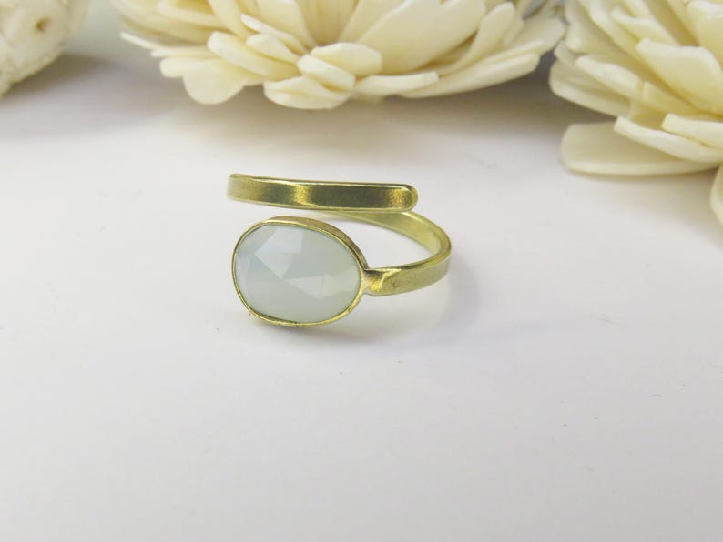 May include: A bypass ring featuring a matte gold-toned metal band and a single oval-shaped, faceted, pale translucent stone set in a bezel. The ring has an open design where the ends of the band do not meet, showcasing a smooth, metallic finish.