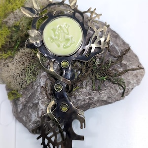 May include: A decorative metal brooch featuring a dark, oxidized finish with an intricate branch-like structure. It includes a central circular pale green cabochon with a frog relief, accented by three small, round, faceted green gemstones.