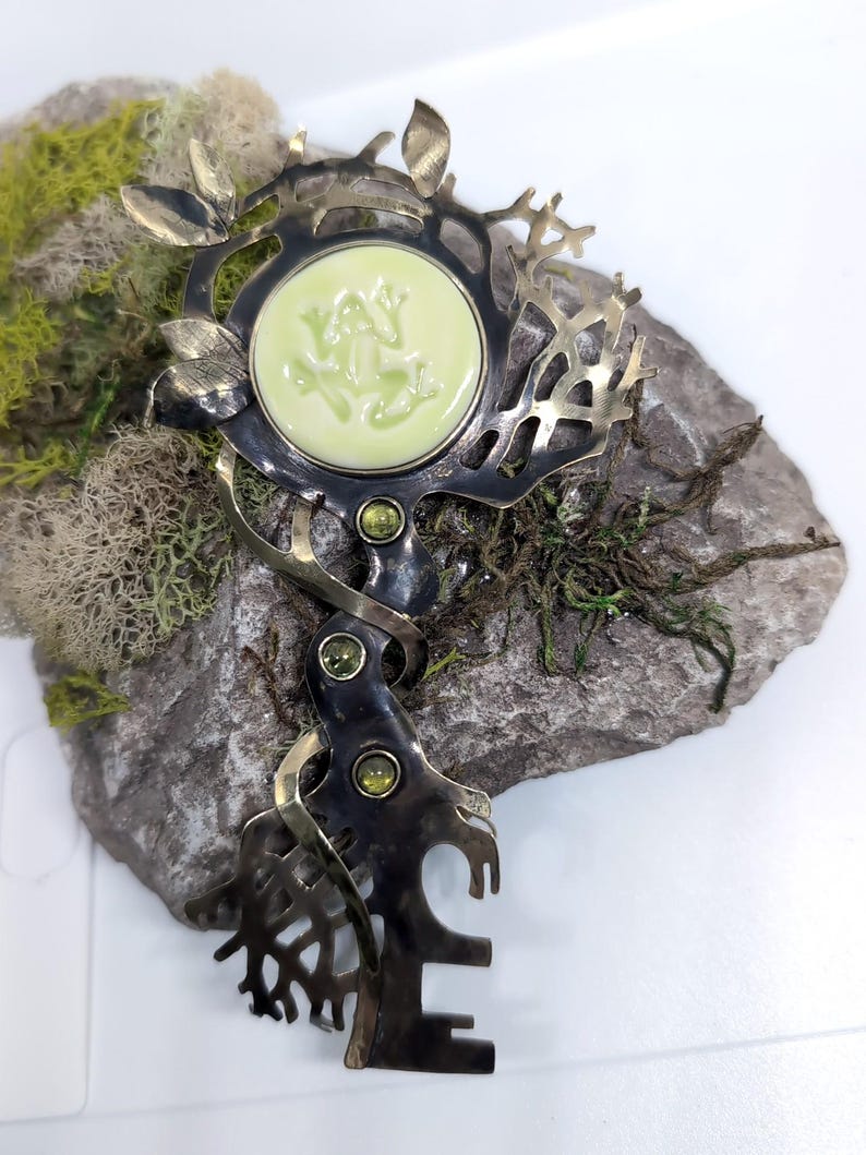 May include: A decorative metal brooch featuring a dark, oxidized finish with an intricate branch-like structure. It includes a central circular pale green cabochon with a frog relief, accented by three small, round, faceted green gemstones.