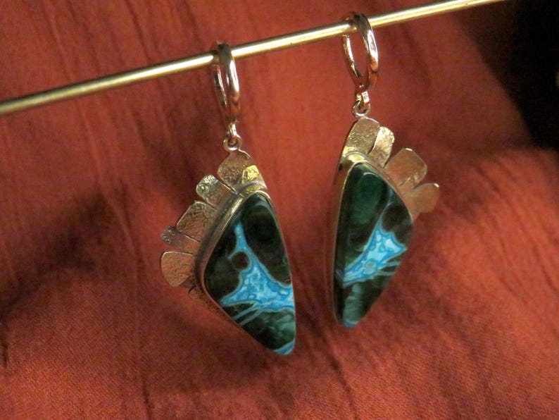 May include: A pair of dangle earrings featuring triangular stones with dark green and bright turquoise veined patterns. The stones are set in silver-toned metal with a textured, fan-like edge detail and secure hinged hoop closures at the top.