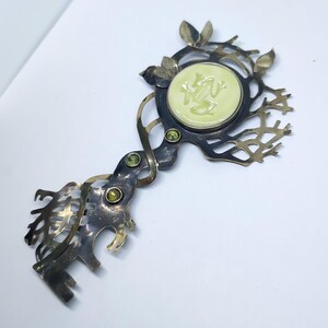 May include: A decorative metal key-shaped brooch featuring a dark oxidized finish with an intricate branch-like design. It includes a central pale yellow circular inlay with a relief of a frog, accented by three small round green gemstones.