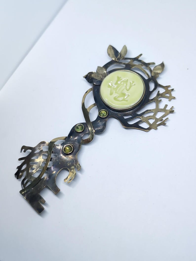 May include: A decorative metal key-shaped brooch featuring a dark oxidized finish with an intricate branch-like design. It includes a central pale yellow circular inlay with a relief of a frog, accented by three small round green gemstones.