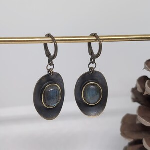 May include: A pair of drop earrings featuring oval-shaped, dark metallic settings with a distressed finish. Each earring contains a central, smooth, iridescent blue-grey stone encased in a thin gold-toned bezel, suspended from a hinged lever-back clasp.