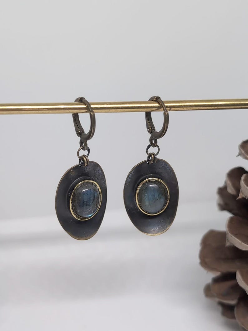 May include: A pair of drop earrings featuring oval-shaped, dark metallic settings with a distressed finish. Each earring contains a central, smooth, iridescent blue-grey stone encased in a thin gold-toned bezel, suspended from a hinged lever-back clasp.