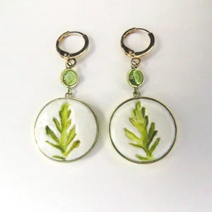 May include: A pair of gold-toned hoop earrings featuring a circular white pendant with a green leaf illustration. Each earring includes a small, round, light green accent stone positioned between the hoop and the larger circular pendant.