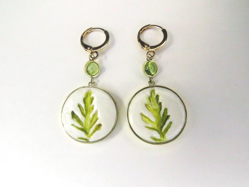 May include: A pair of gold-toned hoop earrings featuring a circular white pendant with a green leaf illustration. Each earring includes a small, round, light green accent stone positioned between the hoop and the larger circular pendant.