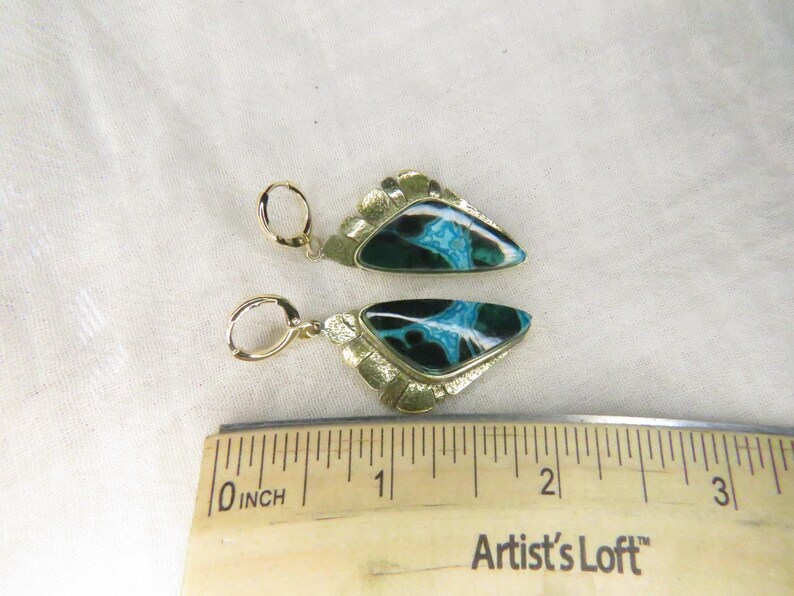 May include: A pair of gold-tone earrings featuring triangular inlays with black and turquoise marbled patterns. The earrings are positioned above a wooden ruler, showing each piece measures approximately 1.25 inches in total length.